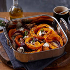 Roasted pumpkin and vegetable dish in a 3-ply stainless steel roasting pan on a wooden table.