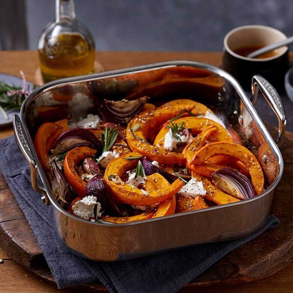 Roasted pumpkin and vegetable dish in a 3-ply stainless steel roasting pan on a wooden table.