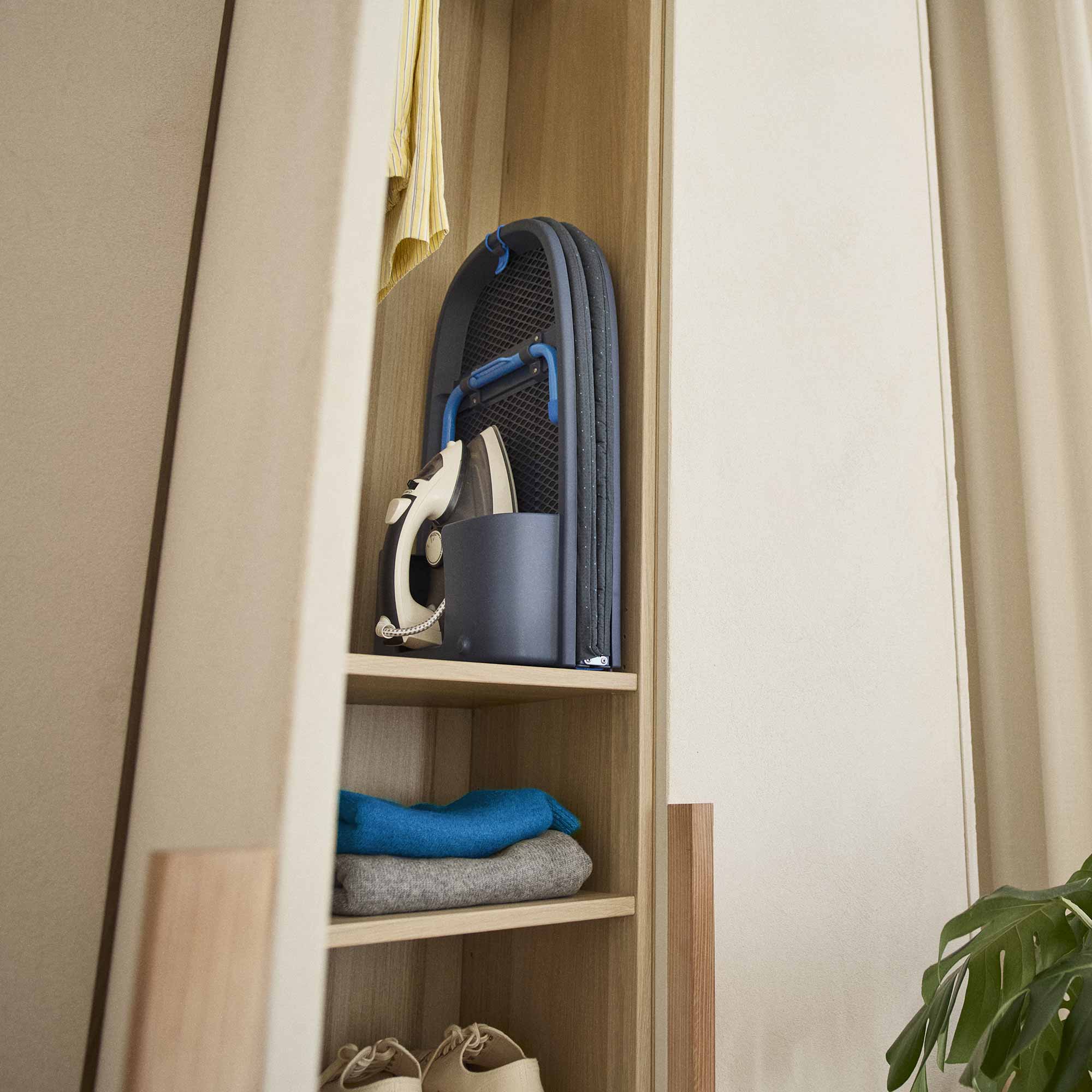 Wooden shelf with folded clothes and a foldable ironing board a neutral wall.