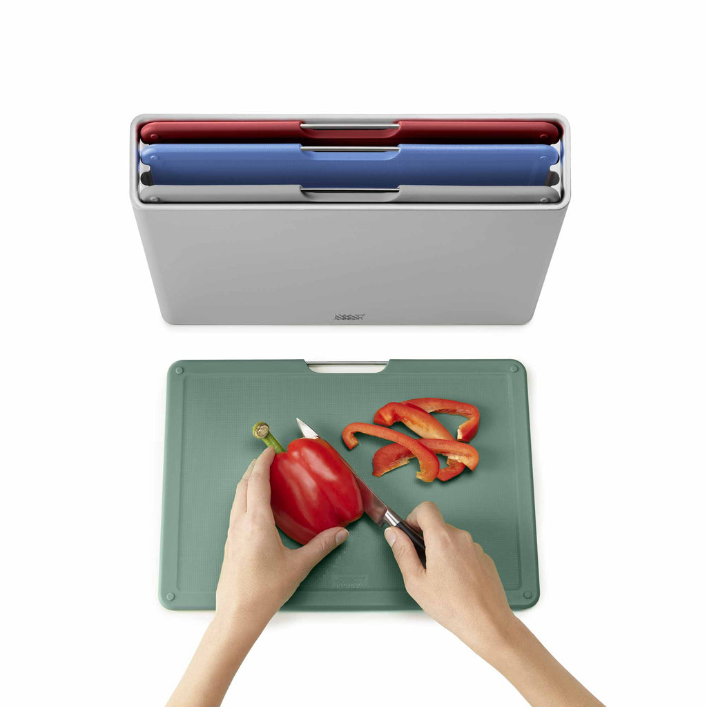 Person cutting a red pepper on a green cutting board with a knife, next to a gray container with colorful compartments.