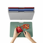 Person cutting a red pepper on a green cutting board with a knife, next to a gray container with colorful compartments.