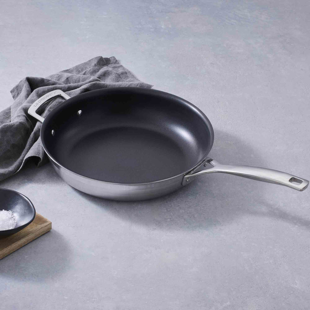 Le Creuset 3-ply Stainless Steel Non-Stick Frying Pan with Helper Handle on grey background with grey cloth