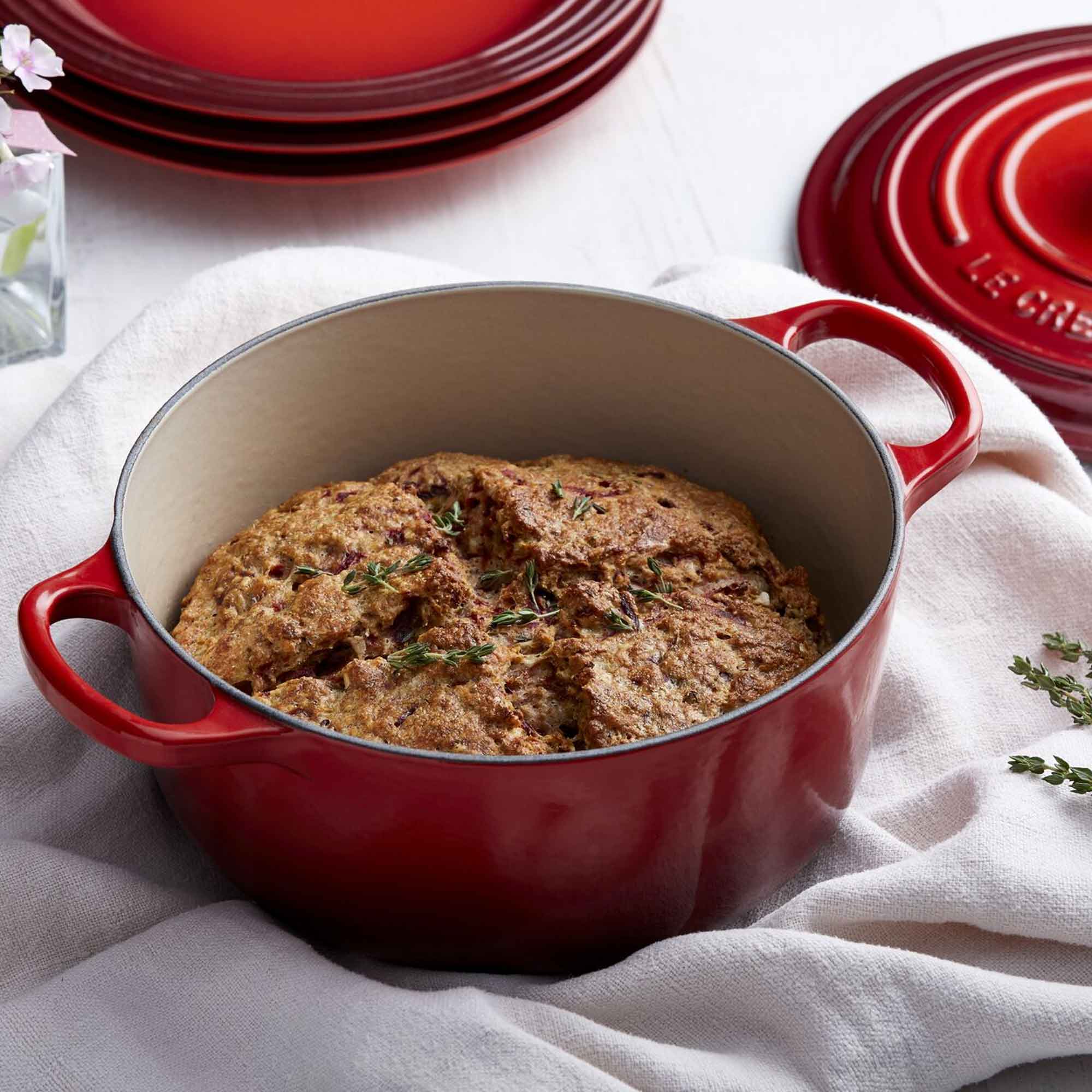 Red cast iron dish with bread and thyme on a white cloth, with Le Creuset branding visible.