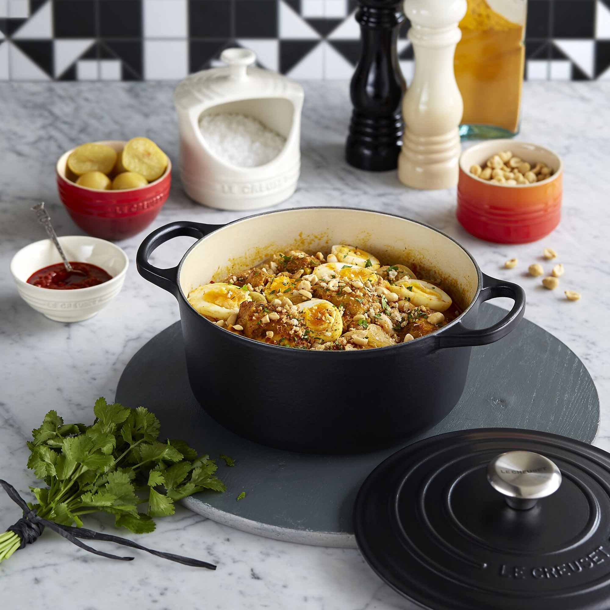Black cast iron pot with food on a marble surface, surrounded by ingredients and utensils.
