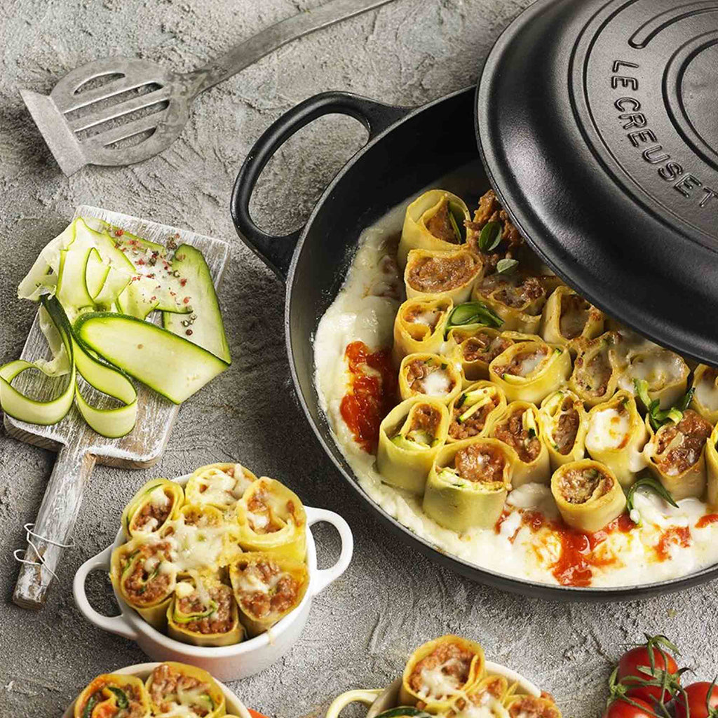 Stuffed pasta rolls in a Le Creuset dish on a gray surface with zucchini and a spatula.