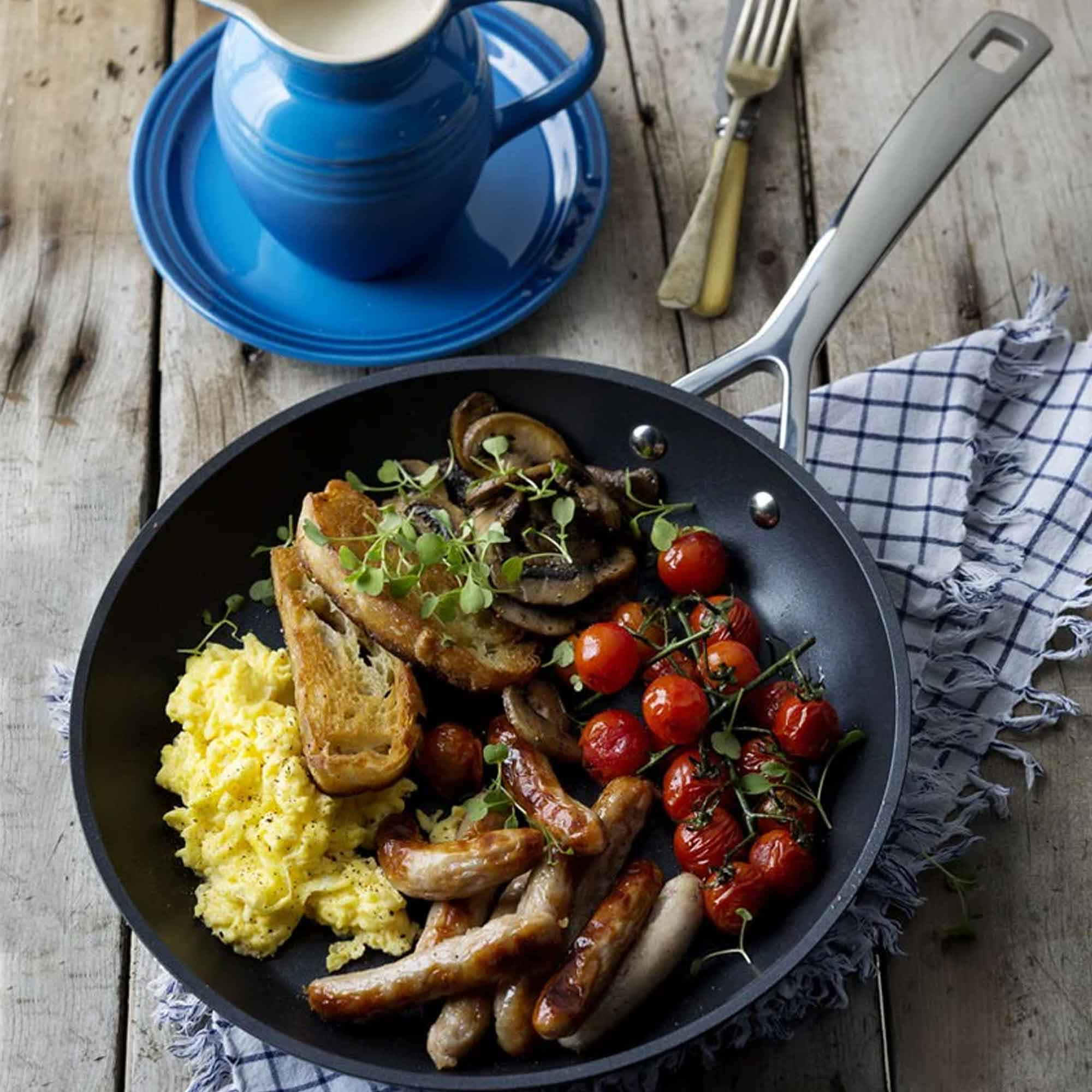 Breakfast dish with sausages, eggs, and roasted tomatoes in a pan on a wooden table.