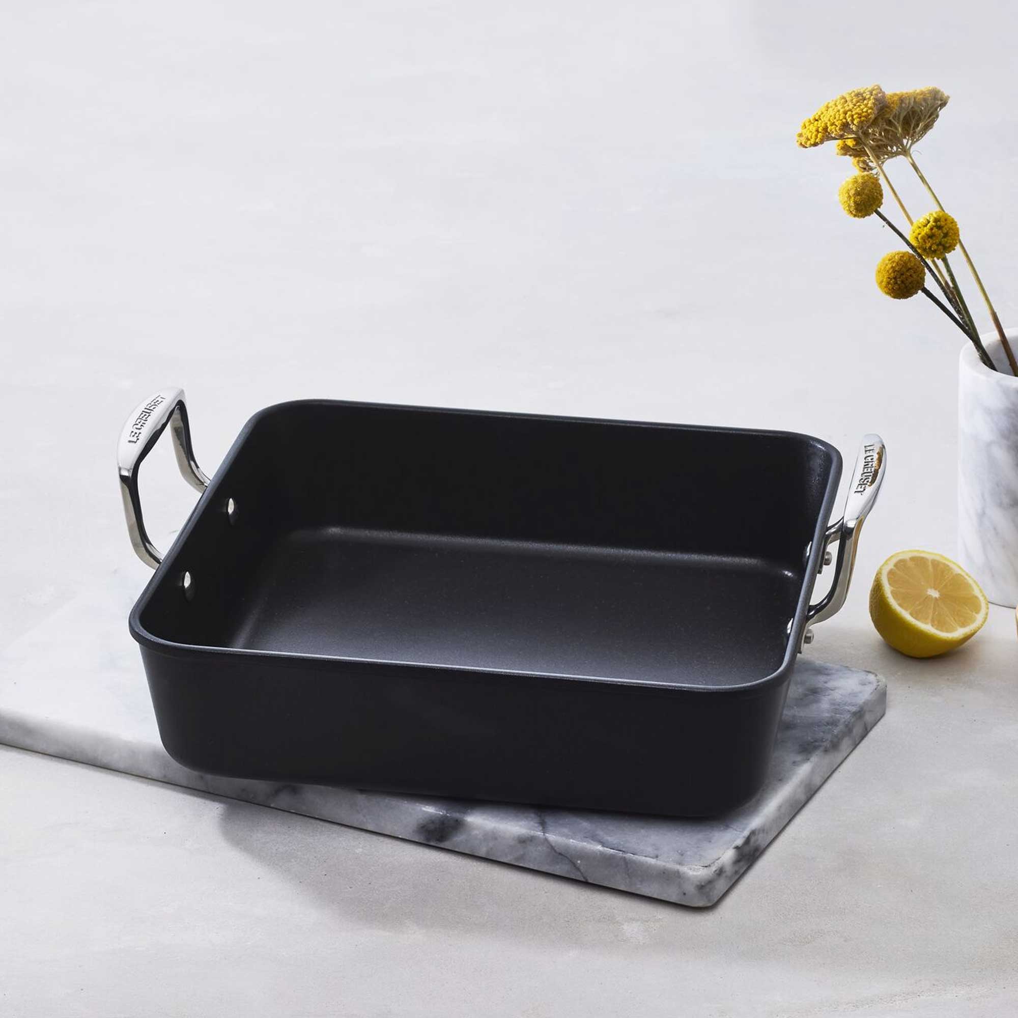 Square roasting pan with handles on a marble surface with a lemon and flowers in the background.
