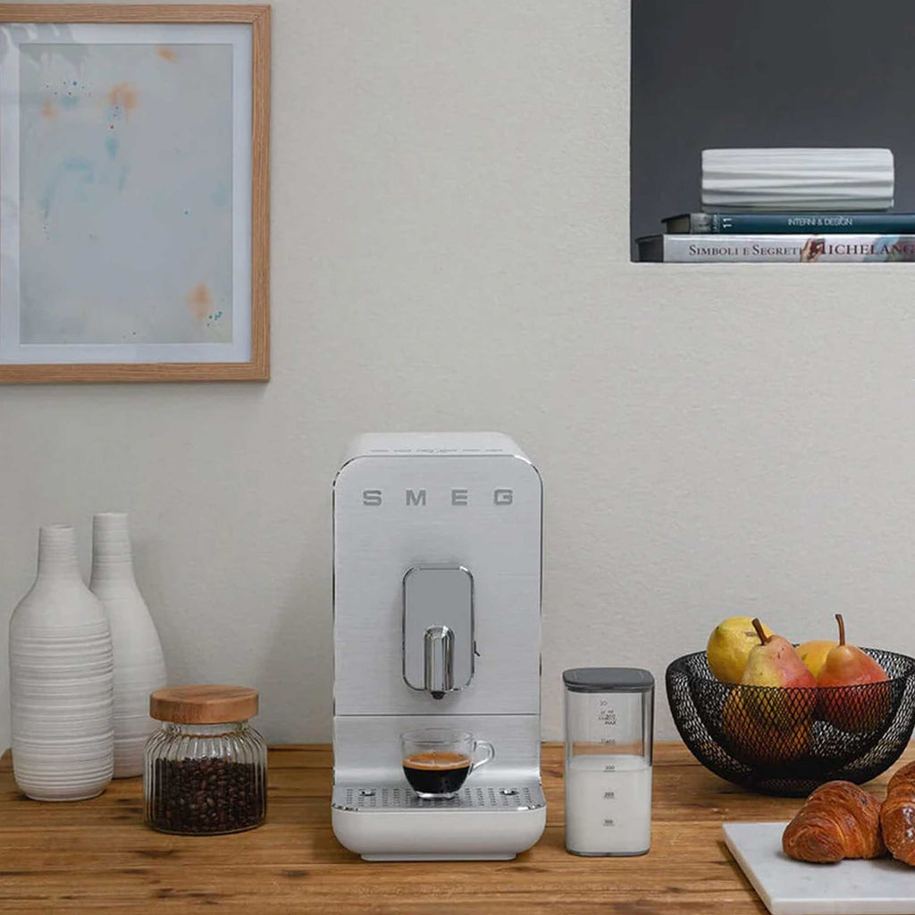 SMEG coffee machine on a wooden surface with a milk container, fruit in a bowl and croissant on a white board