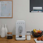 SMEG coffee machine on a wooden surface with a milk container, fruit in a bowl and croissant on a white board