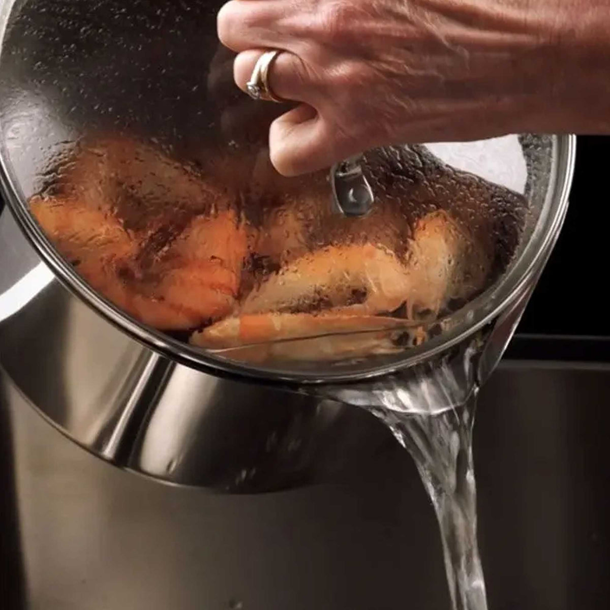 A stainless steel pot with a clear glass lid being lifted by a hand, with hot water pouring out into a sink. Cooked shrimp are visible inside the pot.