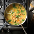 A stainless steel saucepan filled with a chickpea and vegetable soup sits on a gas stovetop. The yellow-orange broth contains chickpeas, green spinach, diced carrots, a lemon slice, and herbs, with a wooden spoon resting inside the pot.