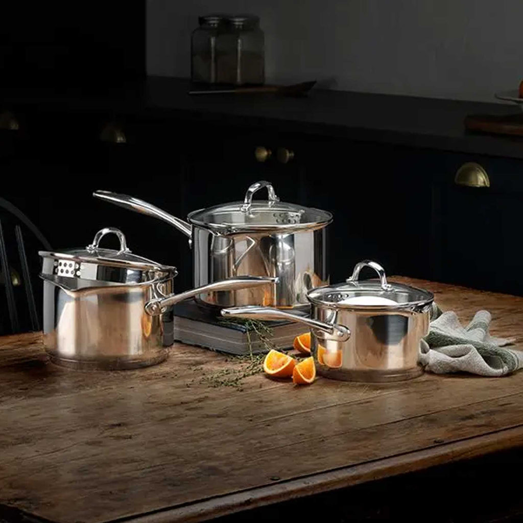 A set of stainless steel cookware, including three pots of varying sizes with glass lids and long handles, arranged on a rustic wooden table in a dark kitchen setting.