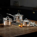 A set of stainless steel cookware, including three pots of varying sizes with glass lids and long handles, arranged on a rustic wooden table in a dark kitchen setting.