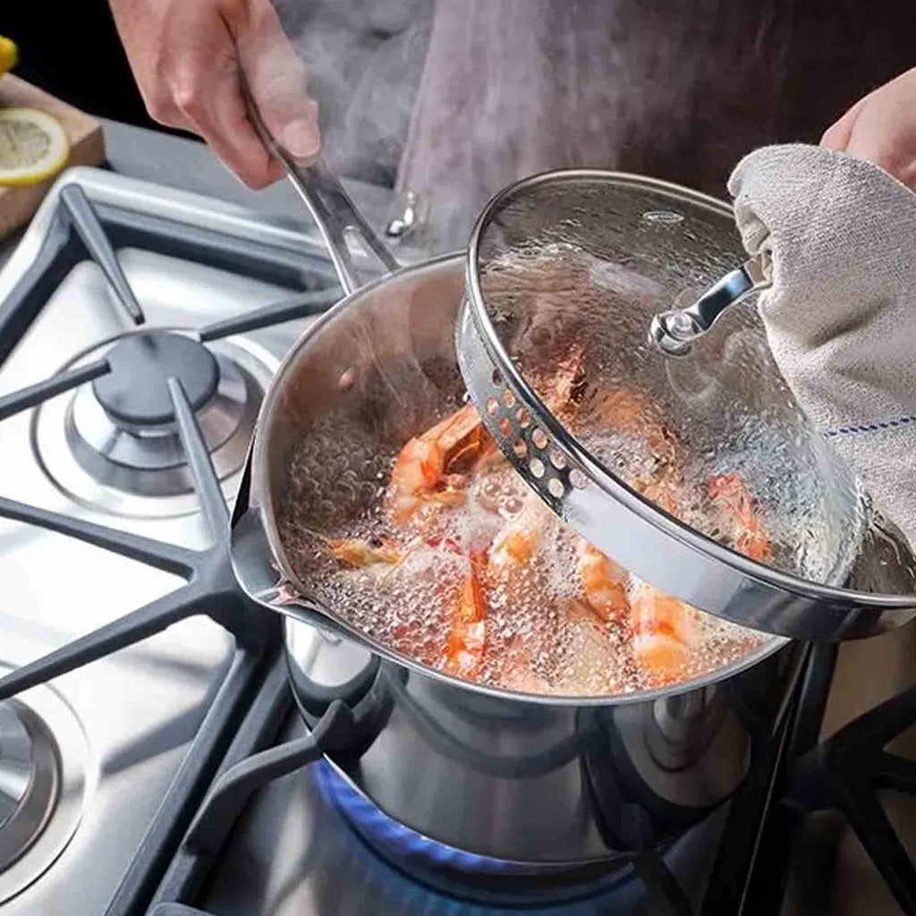 A stainless steel saucepan on a gas stove with shrimp boiling in water, while a person lifts a glass lid using a towel handle. The scene is set in a real kitchen environment.
