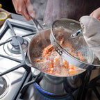 A stainless steel saucepan on a gas stove with shrimp boiling in water, while a person lifts a glass lid using a towel handle. The scene is set in a real kitchen environment.