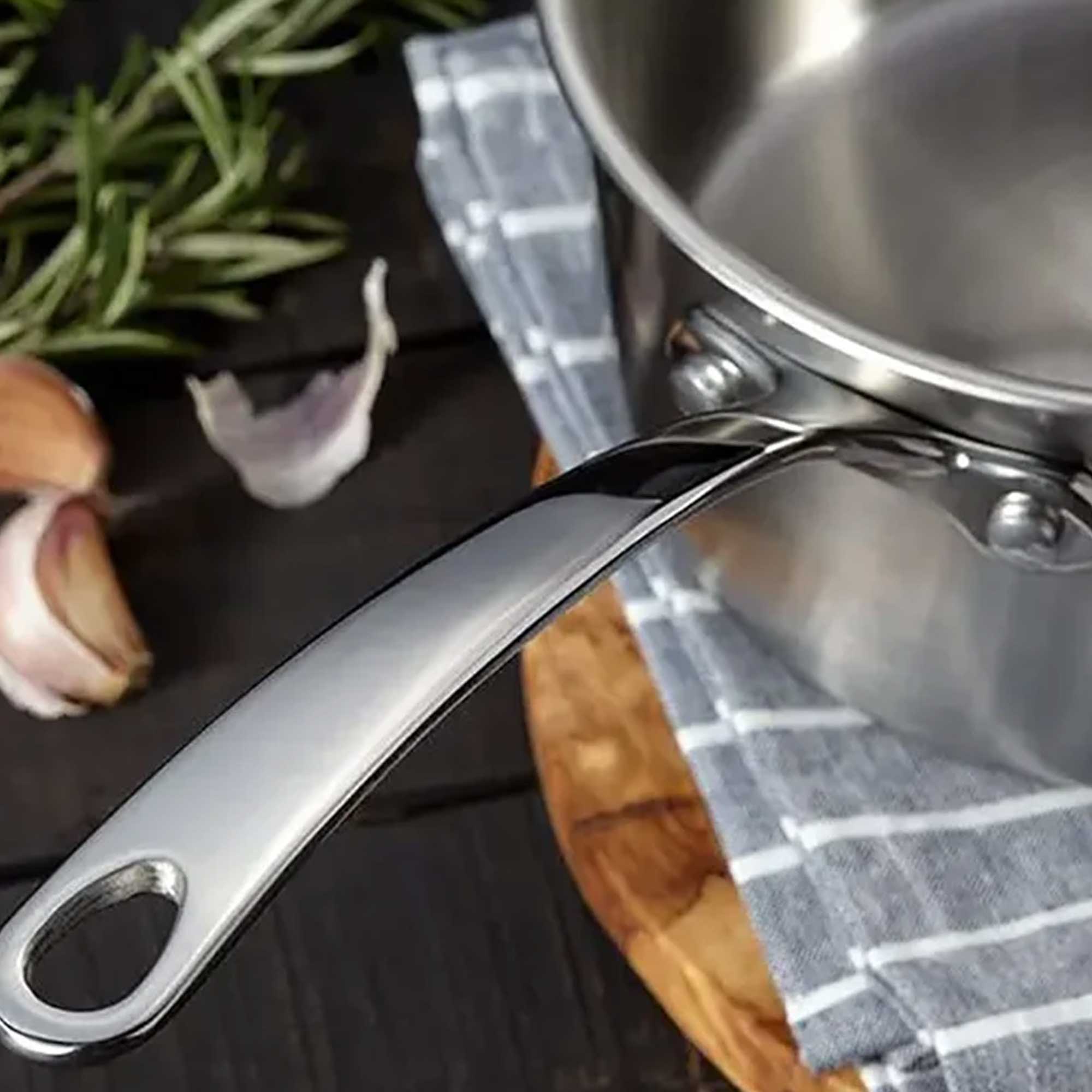 Stainless steel pot with handle on a wooden surface with herbs and garlic.