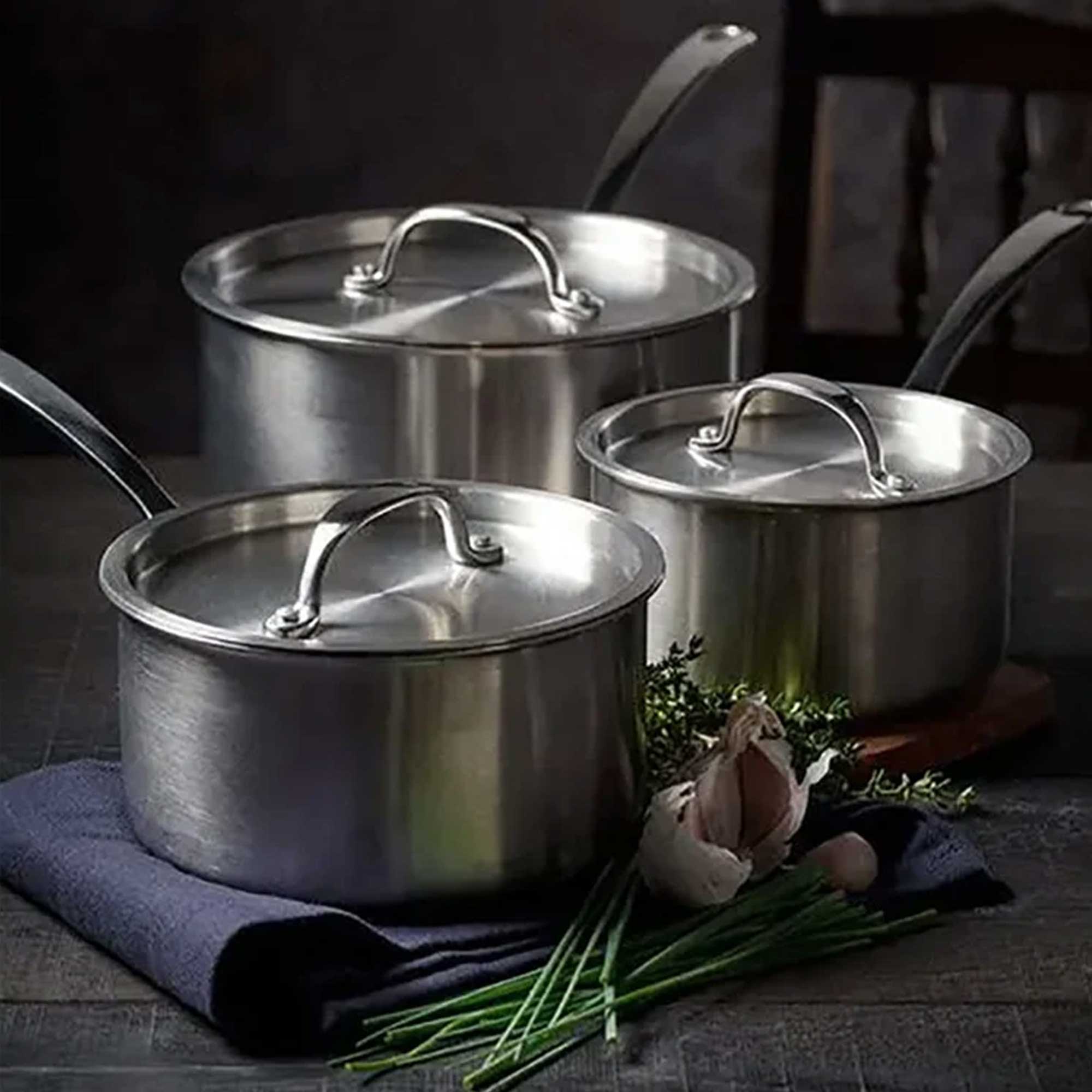 Set of stainless steel cookware with lids on a dark surface with herbs and garlic.