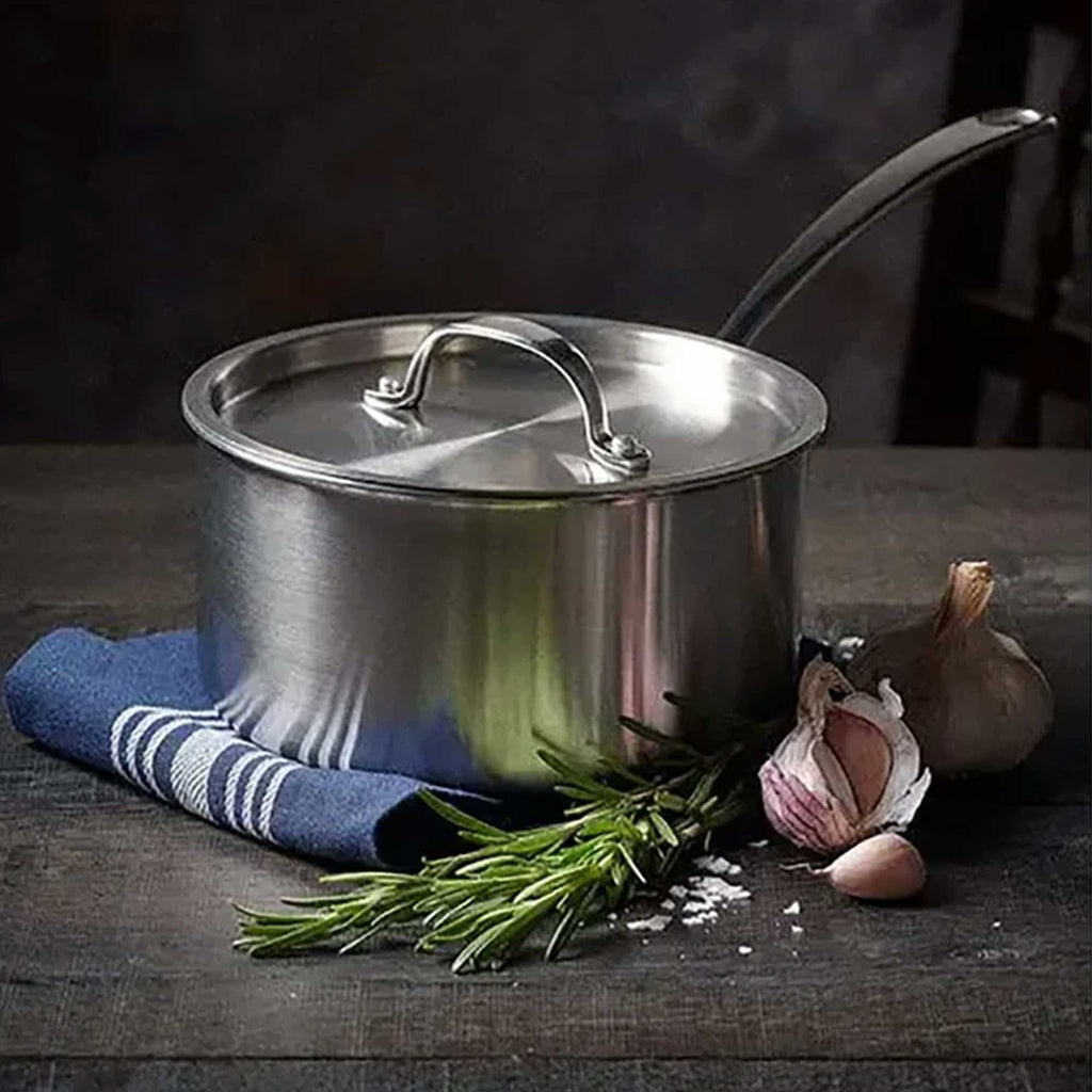 Stainless steel pot with a lid on a wooden surface with herbs and garlic.