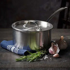 Stainless steel pot with a lid on a wooden surface with herbs and garlic.