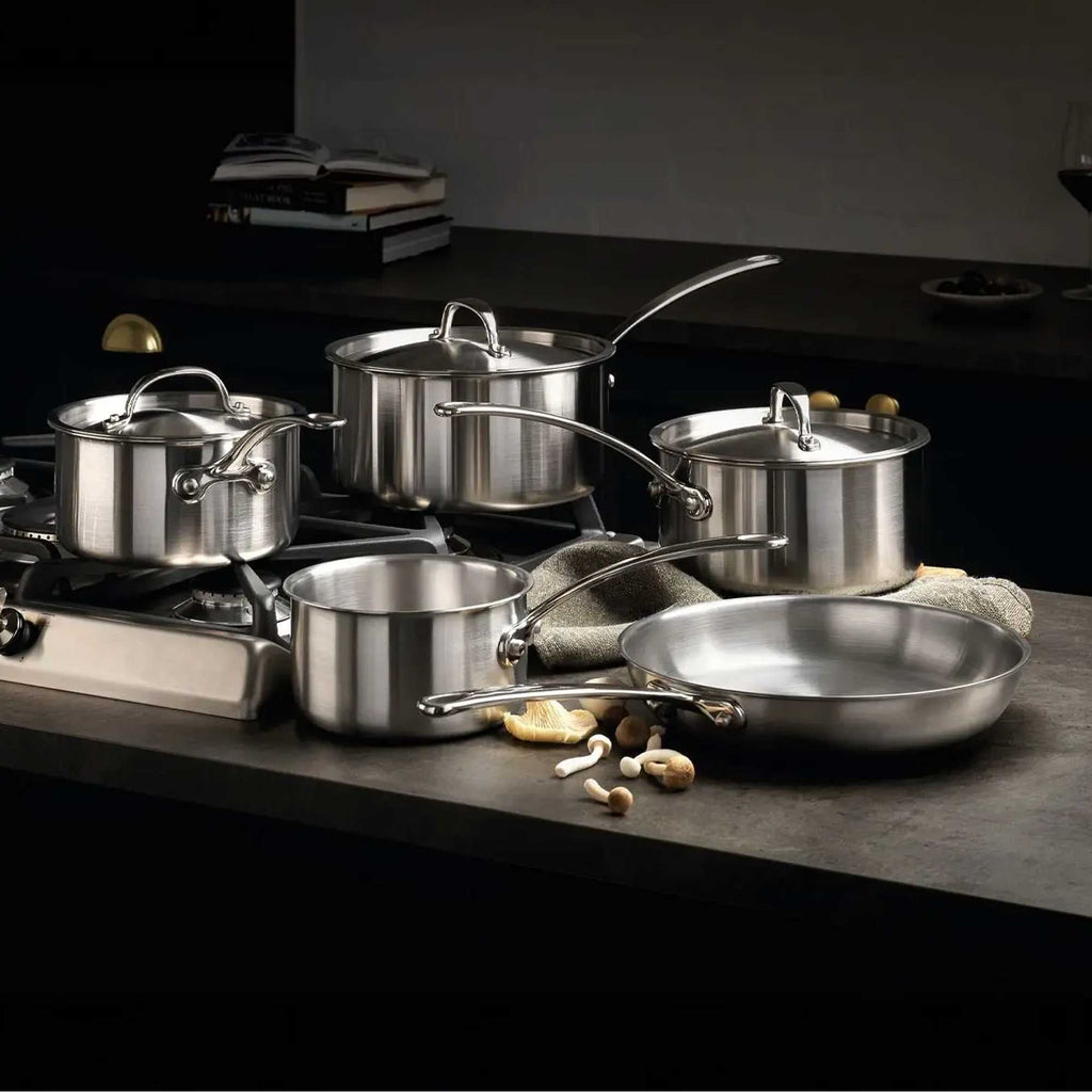A 5-piece stainless steel cookware set with 2 pans sat on a hob and the other 3 on a grey work top with a tea towel and mushrooms