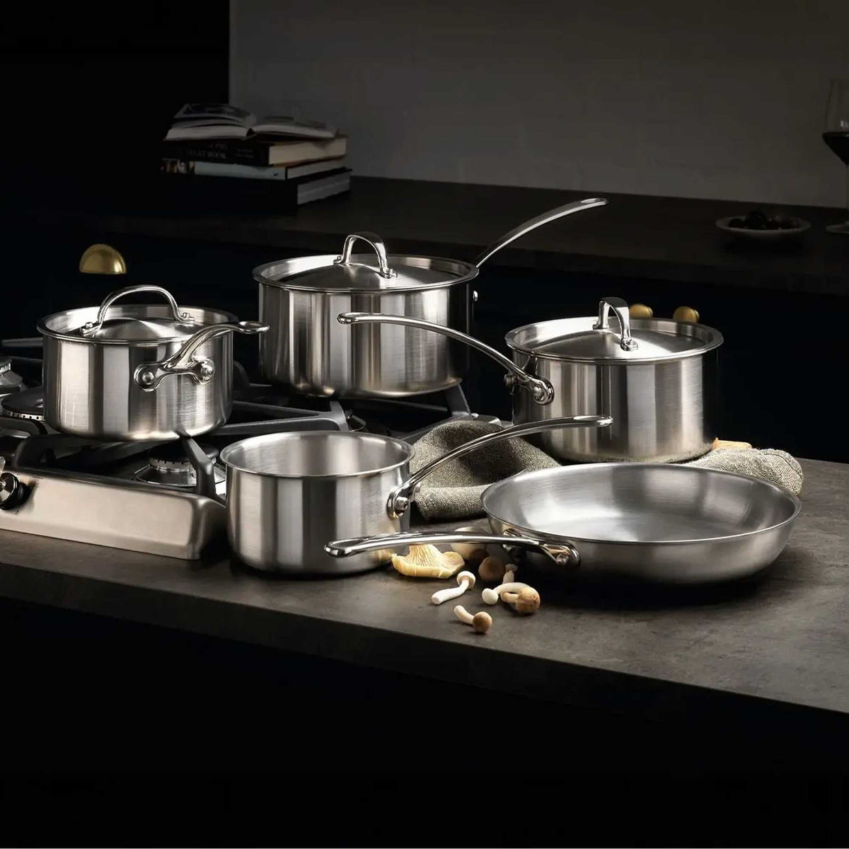 A 5-piece stainless steel cookware set with 2 pans sat on a hob and the other 3 on a grey work top with a tea towel and mushrooms