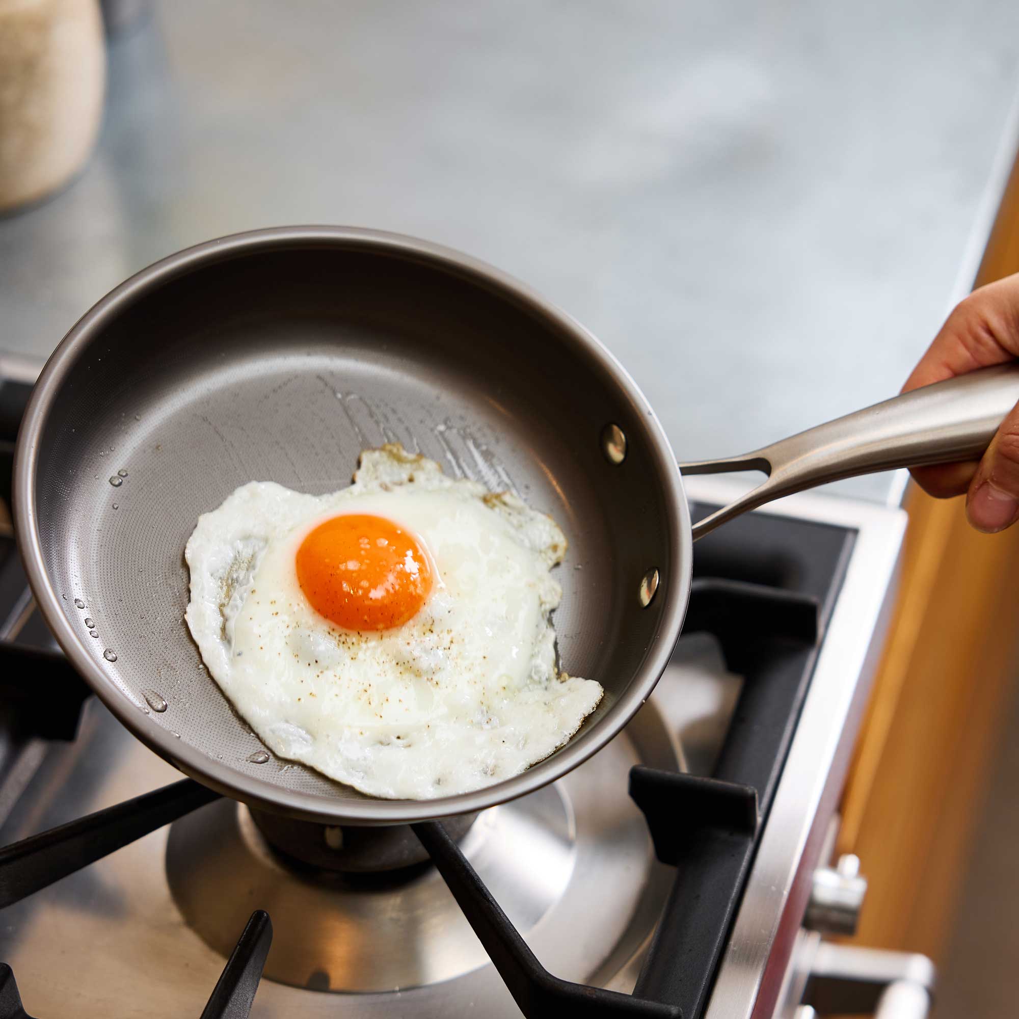 A hand holds a stainless steel frying pan on a gas stovetop, cooking a sunny-side-up egg. The pan has a dark gray non-stick interior and a silver metal handle.