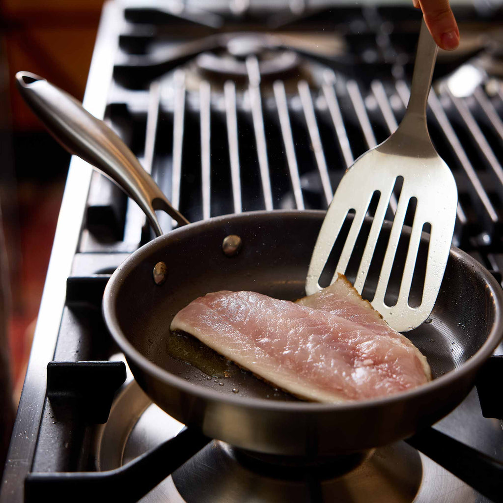 A stainless steel frying pan on a gas stovetop, containing a piece of raw fish being held by a metal spatula.