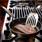 A stainless steel frying pan on a gas stovetop, containing a piece of raw fish being held by a metal spatula.