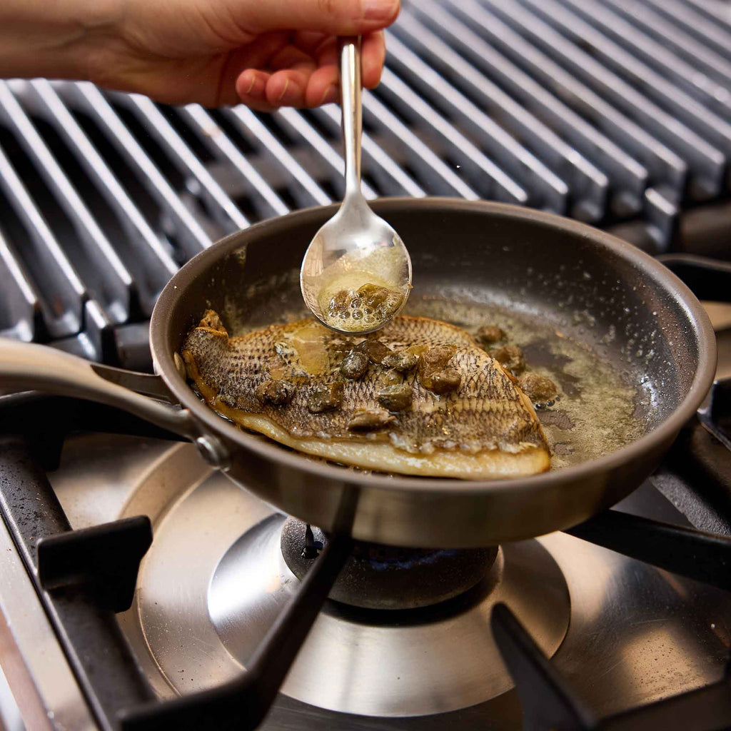 A close-up of a fish cooking in a dark pan on a gas stovetop, with a hand holding a spoon and pouring liquid over the fish.