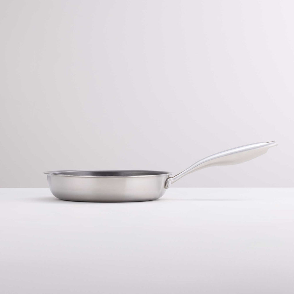 A stainless steel frying pan with a long, curved handle, shown from a side profile on a clean, light gray studio background.