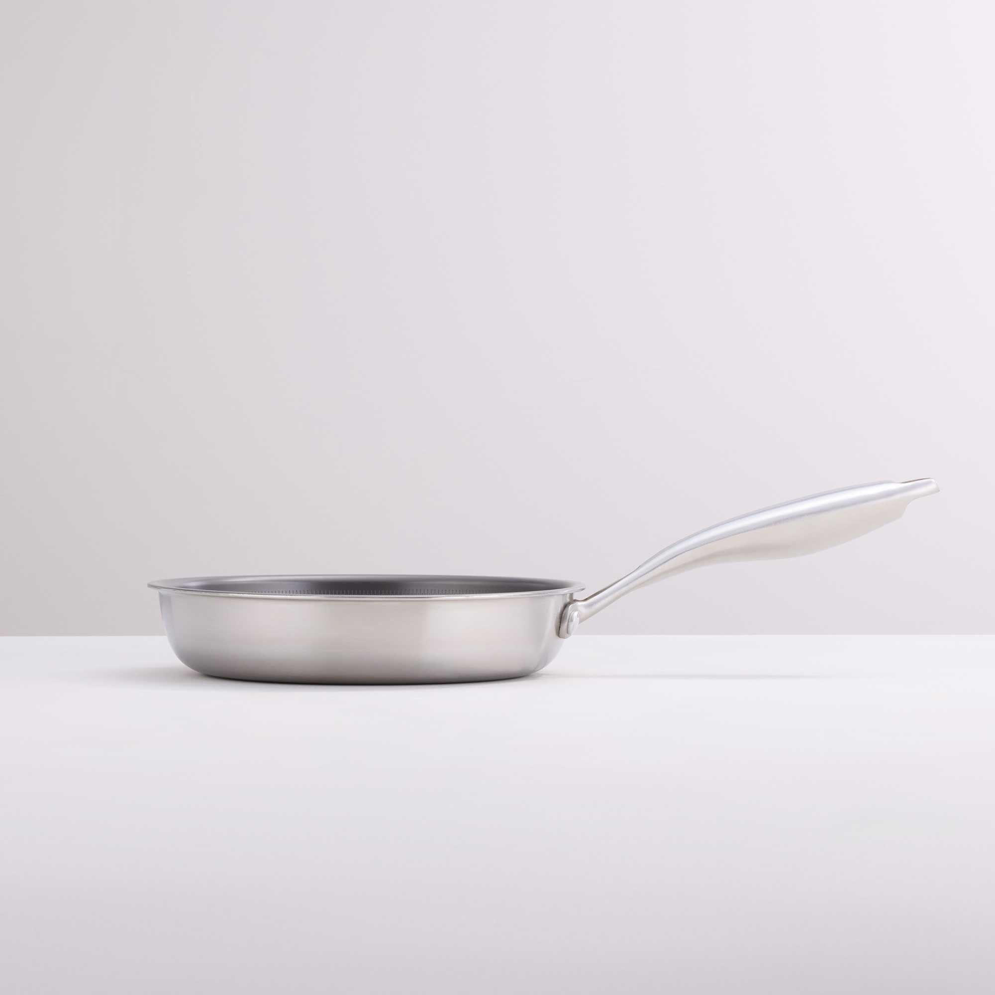 A stainless steel frying pan with a long, curved handle, shown from a side profile on a clean, light gray studio background.