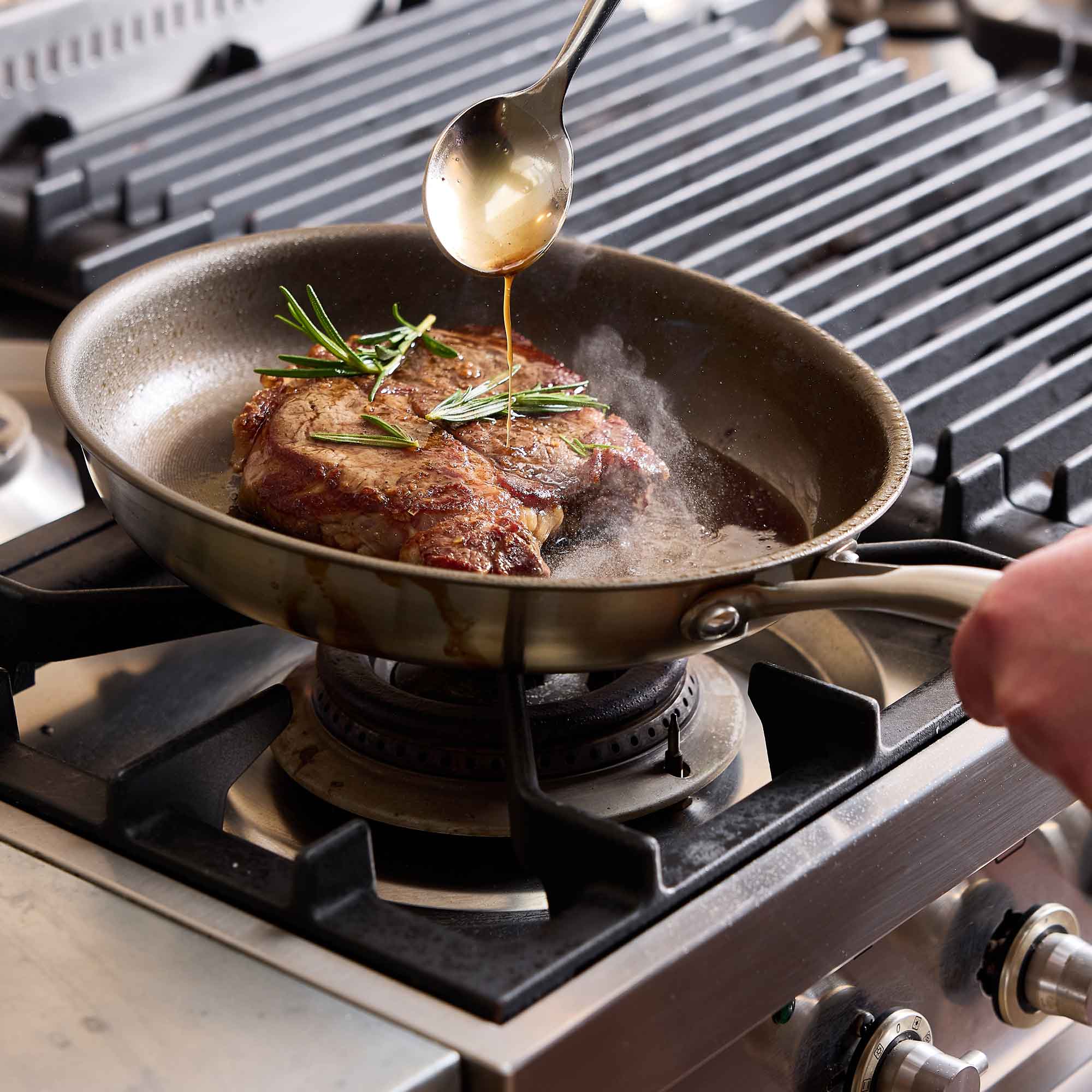 A stainless steel frying pan on a gas stove burner, showing a seared steak with rosemary sprigs and a spoon pouring sauce over it. A person's hand is visible holding the pan handle.