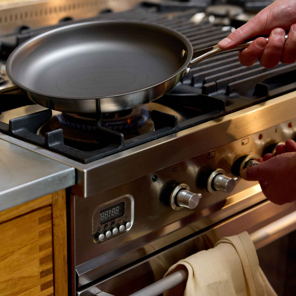 A stainless steel frying pan with a dark non-stick coating sits on a gas stovetop, with a person's hand holding the handle and adjusting the stove knobs.