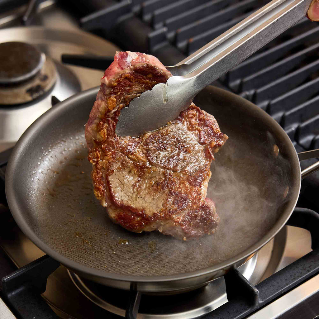 A seared steak is being lifted with metal tongs out of a dark frying pan on a gas stovetop.