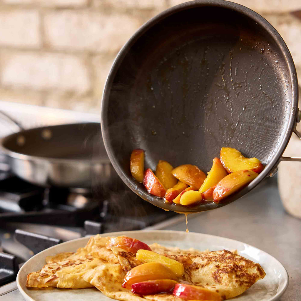 A dark nonstick frying pan pouring cooked peach slices over an folded crepe on a plate, with a stovetop and second pan blurred in the background.
