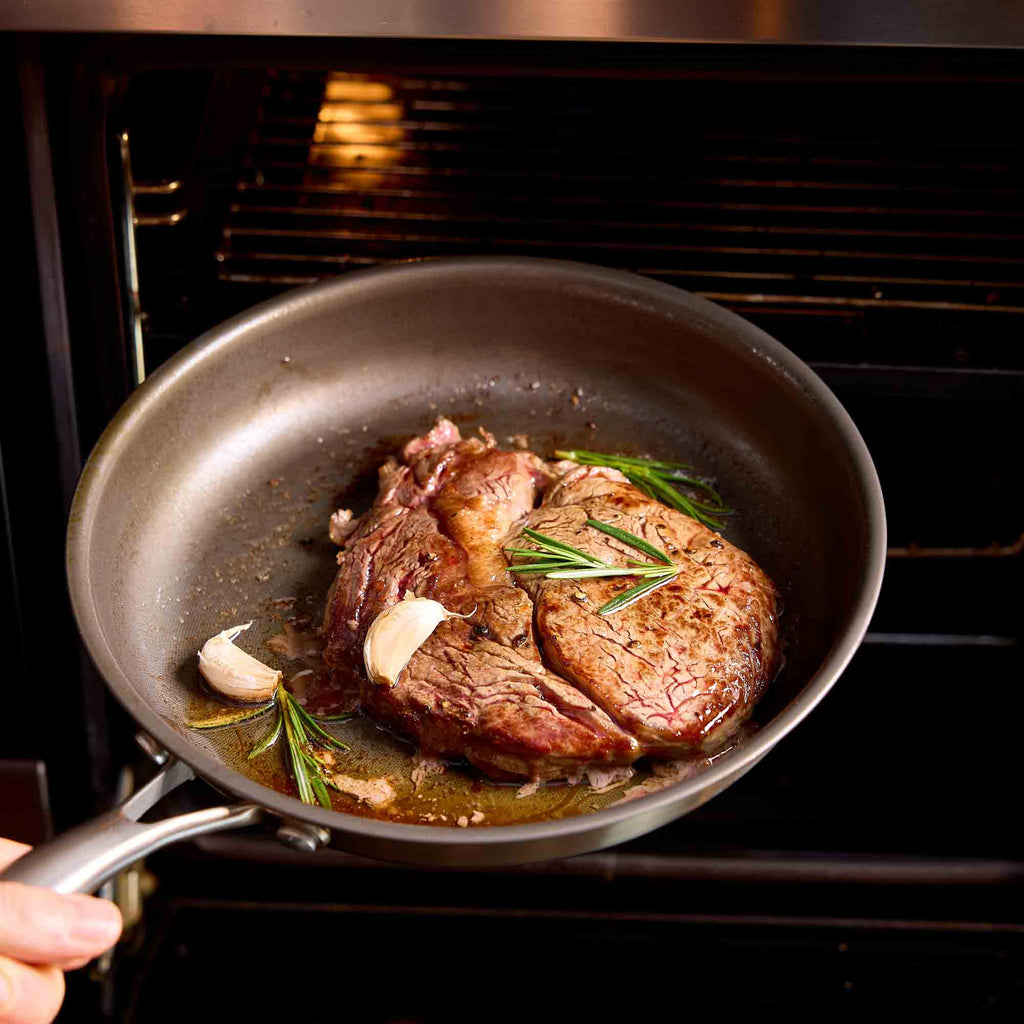 A dark non-stick frying pan on a stovetop with a seared steak, garlic, and rosemary, with a hand holding the handle as it is being pulled into an oven.