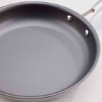 A close-up of a dark gray non-stick frying pan with a silver metal handle and rivets, set against a light off-white background.