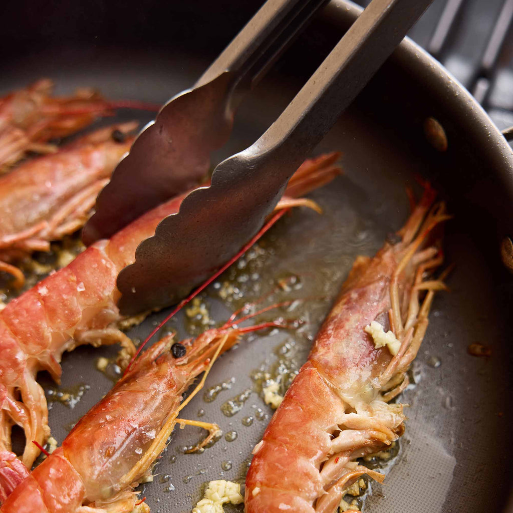 Close-up of cooked prawns frying in a dark pan with metal tongs resting on top.