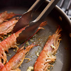 Close-up of cooked prawns frying in a dark pan with metal tongs resting on top.
