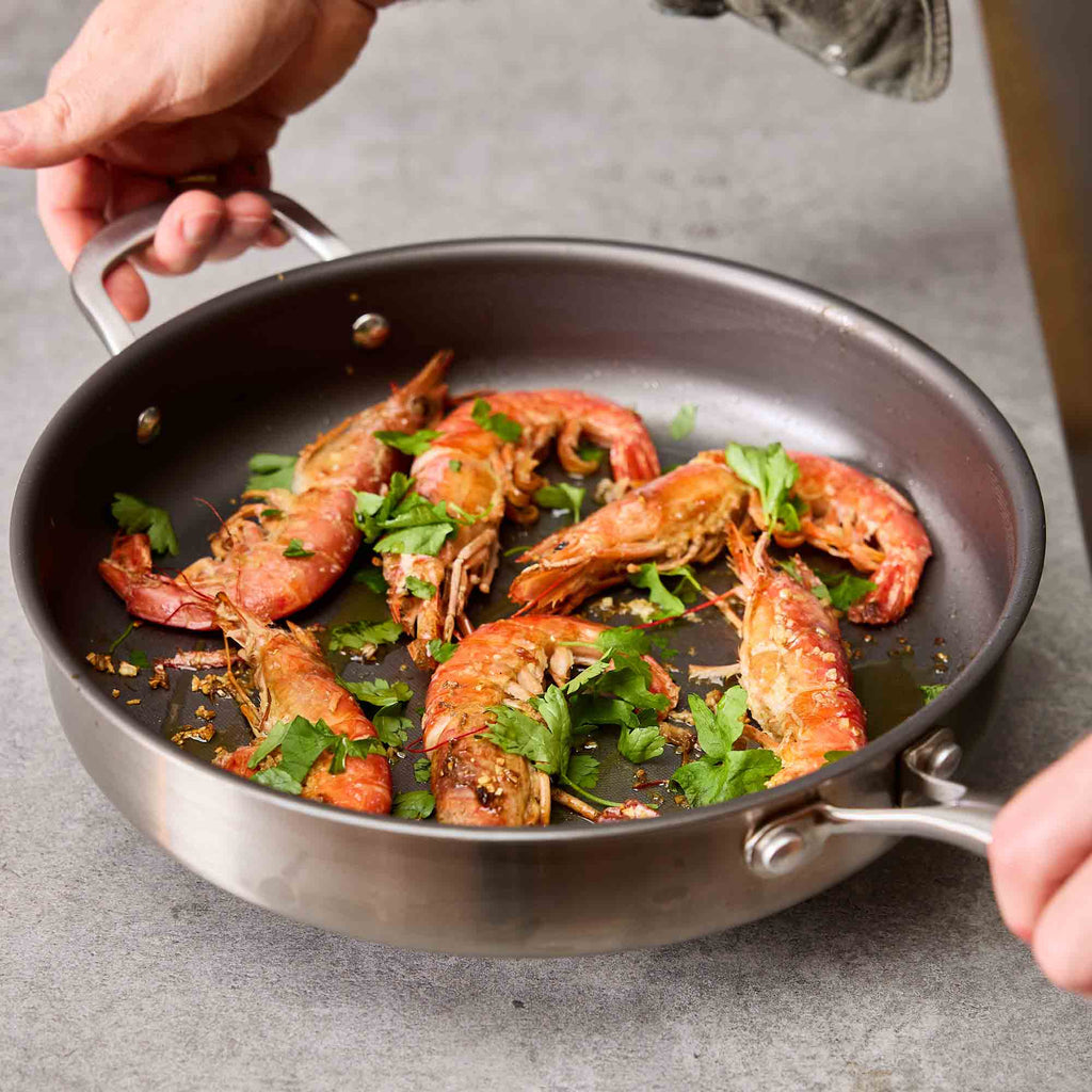 A stainless steel saute pan with two handles containing cooked shrimp topped with chopped green herbs, held by hands over a gray countertop.