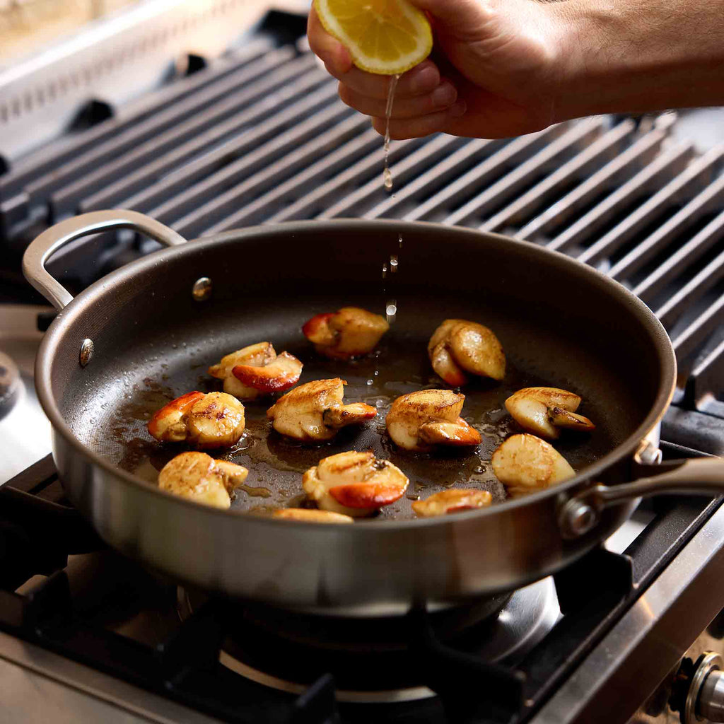 A stainless steel sauté pan on a gas stovetop, containing seared shrimp, with a hand squeezing lemon juice over the pan.