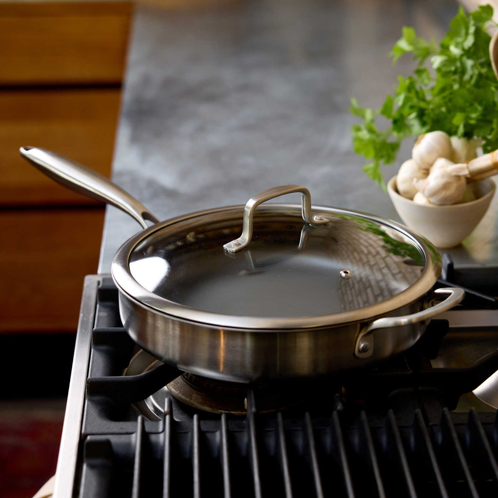A stainless steel sauté pan with a glass lid sits on a gas stovetop in a kitchen setting, with a bowl of garlic and fresh parsley blurred in the background.