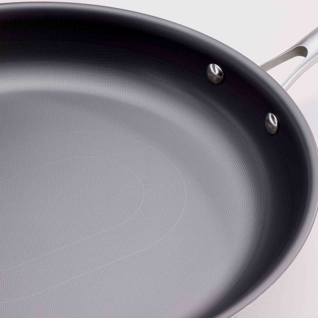 Close-up of a dark non-stick saute pan with a textured interior and a silver metal handle attachment, shown against a light background.