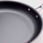 Close-up of a dark non-stick saute pan with a textured interior and a silver metal handle attachment, shown against a light background.