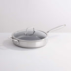 A stainless steel sauté pan with a clear glass lid and long handle, photographed on a clean white studio background.