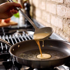 A hand holds a metal ladle pouring a smooth, pale yellow batter into a dark non-stick frying pan on a gas stovetop, with a rustic brick wall in the background.
