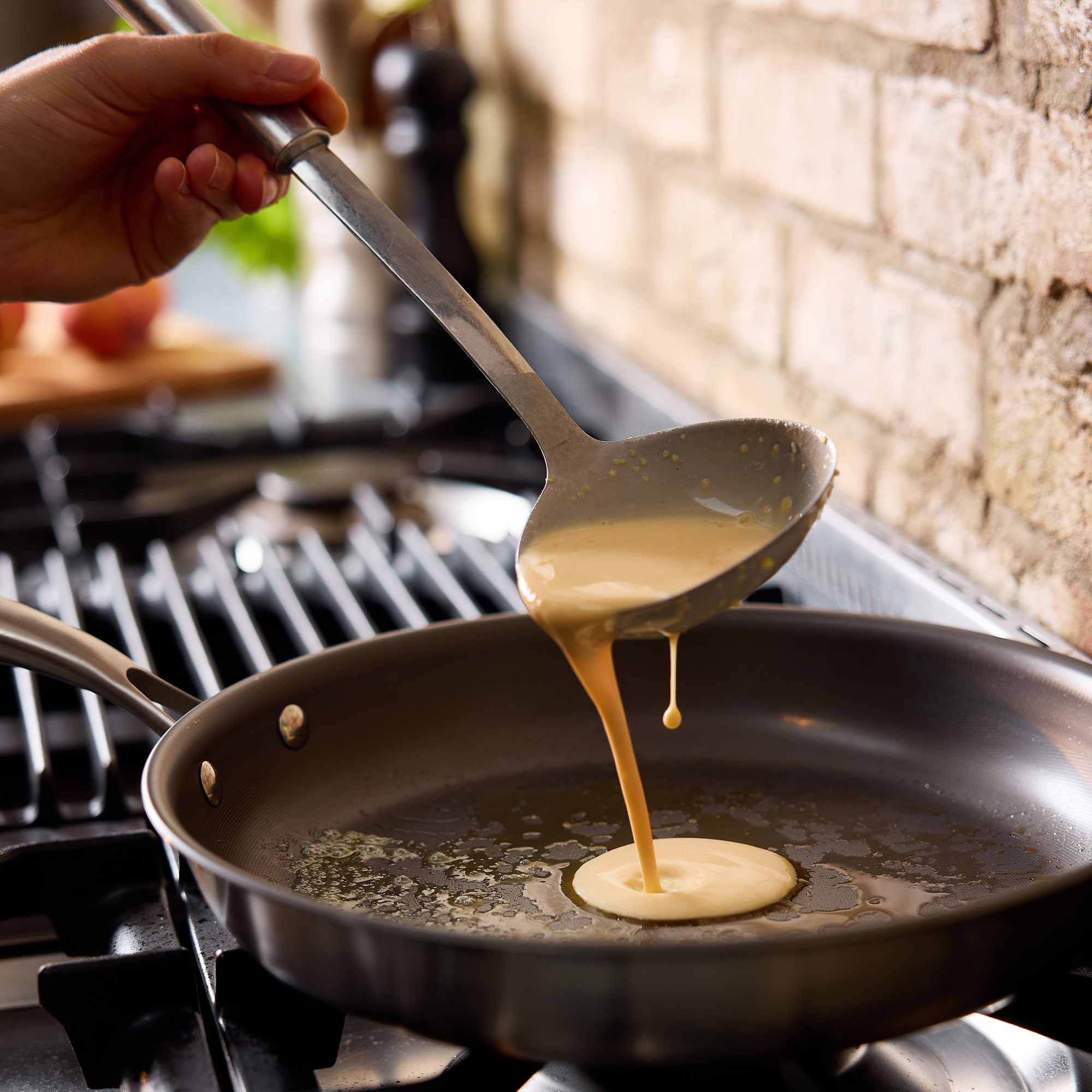 A hand holds a metal ladle pouring a smooth, pale yellow batter into a dark non-stick frying pan on a gas stovetop, with a rustic brick wall in the background.