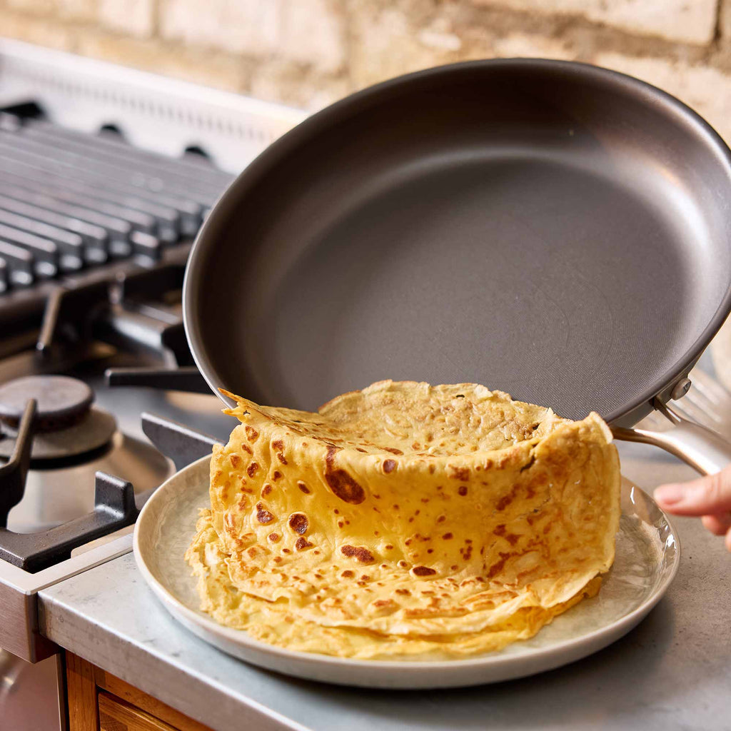 A dark nonstick frying pan is being used to lift a stack of golden-brown crepes from a light gray plate on a stovetop, with a hand holding the pan handle.