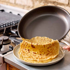 A dark nonstick frying pan is being used to lift a stack of golden-brown crepes from a light gray plate on a stovetop, with a hand holding the pan handle.