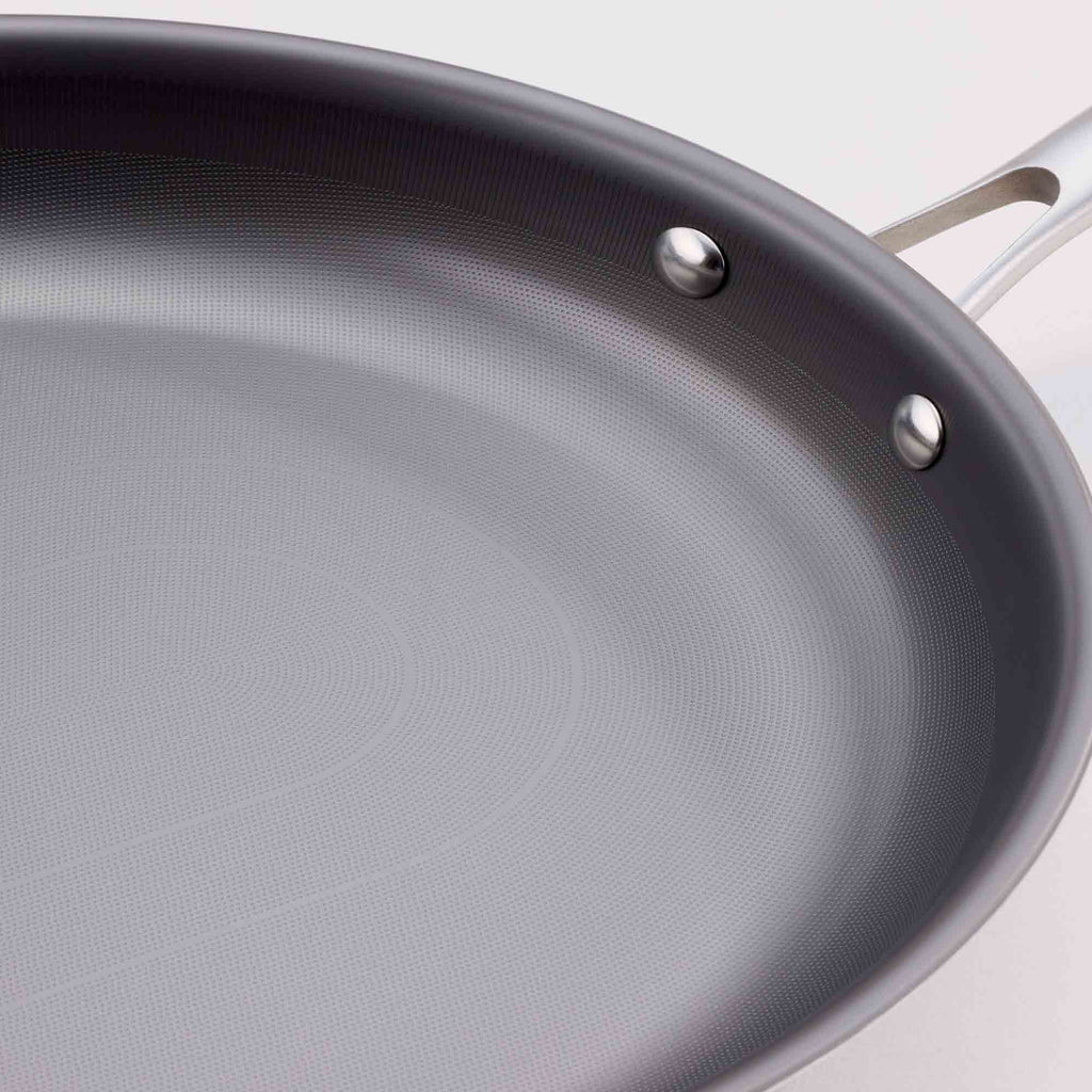 Close-up of a dark gray non-stick frying pan with a textured interior surface and stainless steel rivets, showing part of the silver handle against a light background.